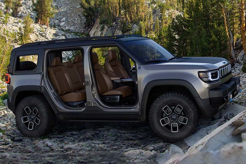 A 2025 Jeep Recon electric vehicle sits at the bottom of a rugged rocky hill with its doors removed, revealing its comfortable, modern interior.