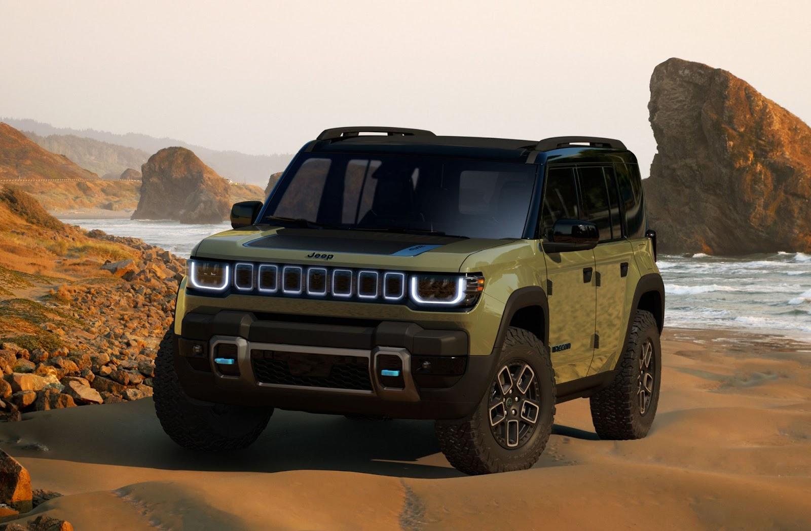 A futuristic Jeep Recon SUV parked on a sandy beach with rocky outcrops behind.