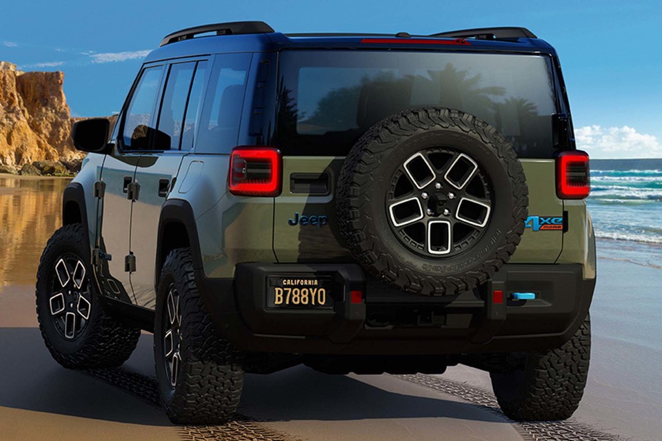 A photo of the rear of an all-electric Jeep Recon on a beautiful beach with cliffs and the surf in the background.